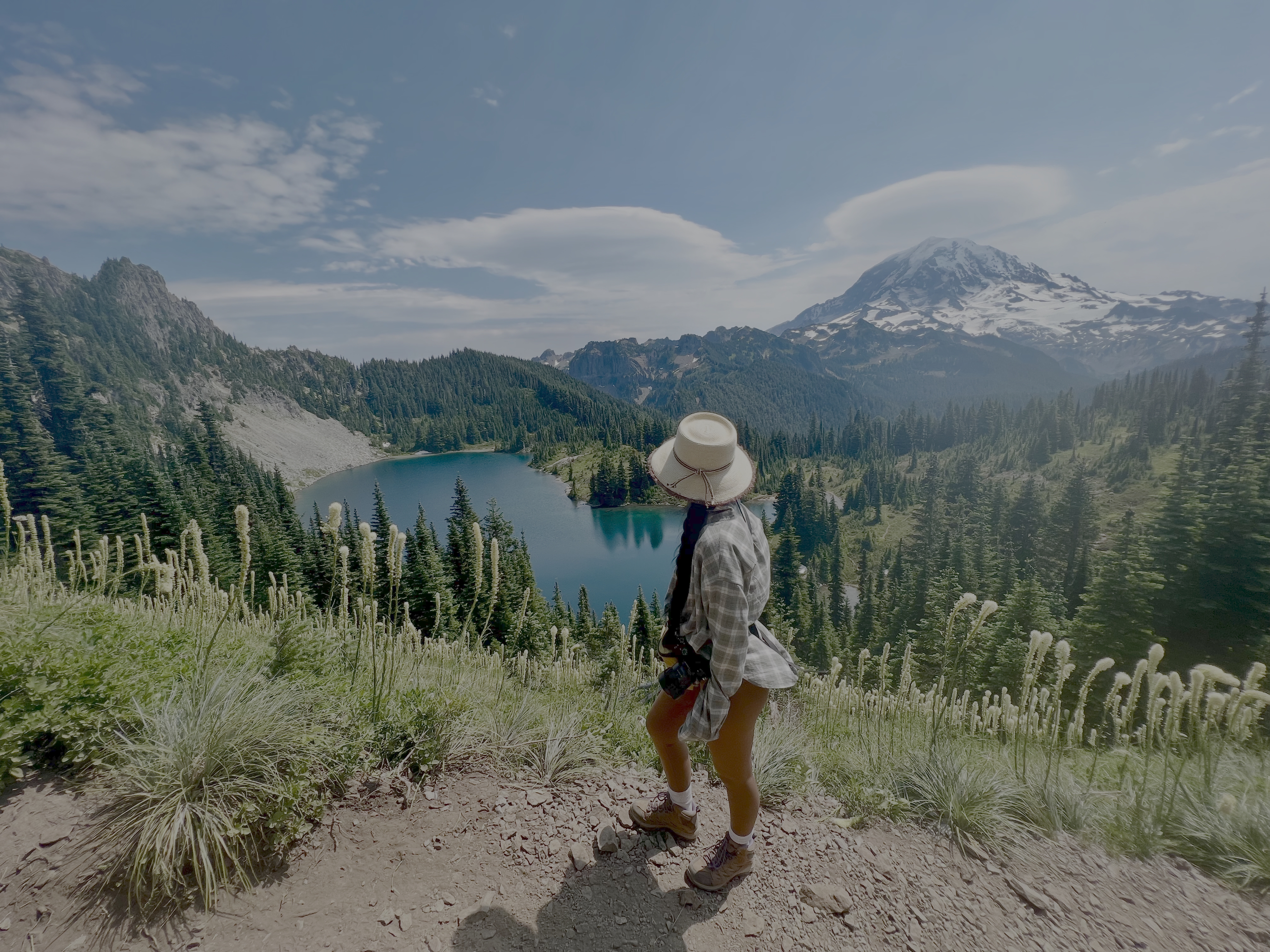 Jenna Funakoshi hiking on a dirt trail above a beautiful view of a lake surrounded by trees, with a snowcapped mountain in the distance. The sky is blue.