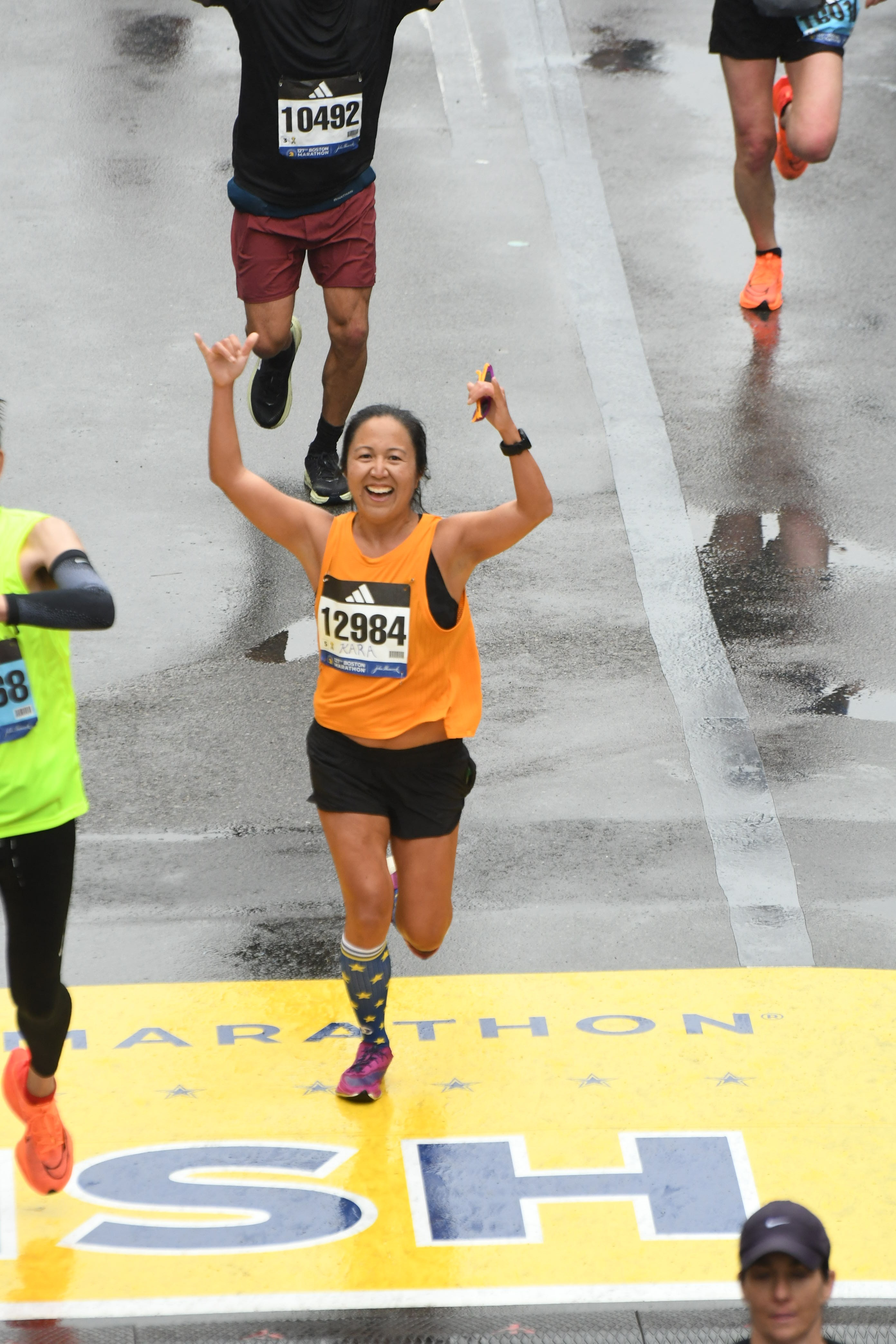 Kara Saiki in an orange and black running outfit at the finish line of the Boston Marathon with her arms raised high in celebration and a big smile.