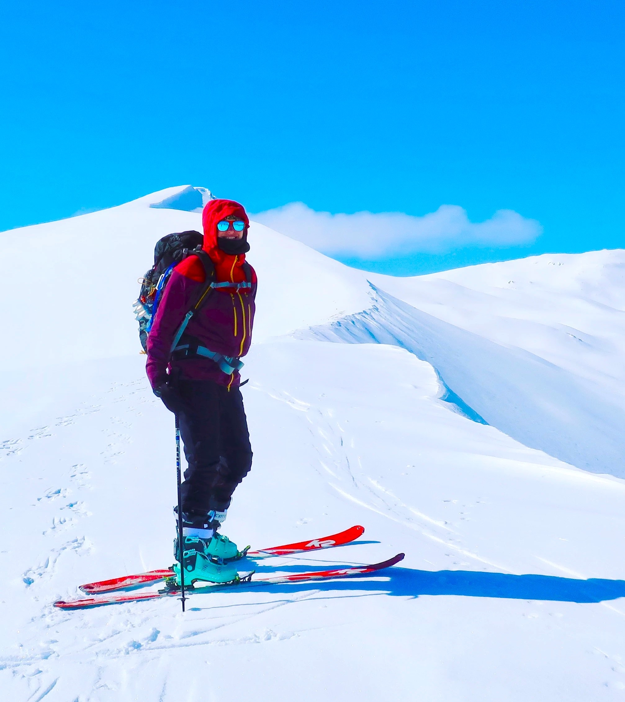 Leah grout on skis on top of a snow covered mountain, fully bundled up with a warm protective gear. There is a backpack on her back. The sky is bright blue.